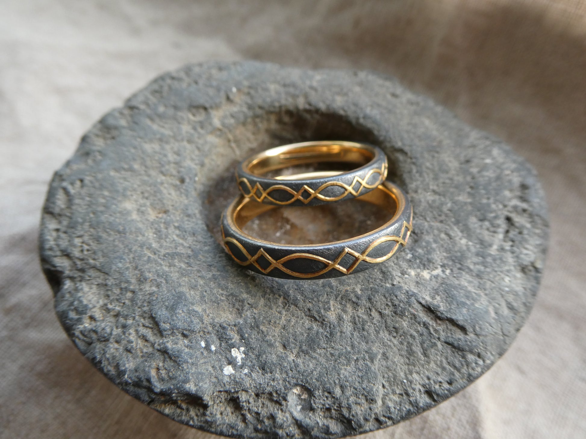 gold inlaid wedding bands with design set against blackened silver displayed on stone
